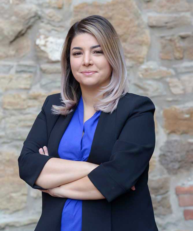 smiling young woman wearing navy blazer and blue shirt with arms crossed in front of stone wall