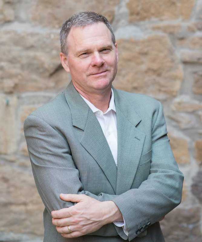 man wearing grey suit with arms crossed in front of stone wall