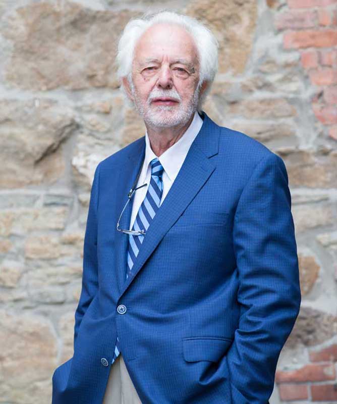 man with white hair and beard wearing blue suit and striped tie in front of stone wall