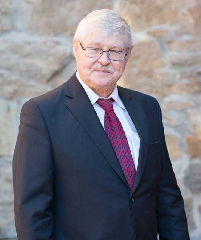 man with grey hair and moustache wearing glasses black suite and tie in front of stone wall