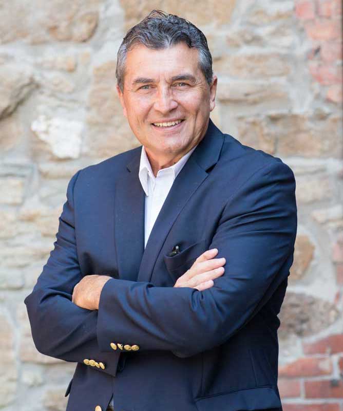 smiling man wearing navy suit with arms crossed in front of stone wall