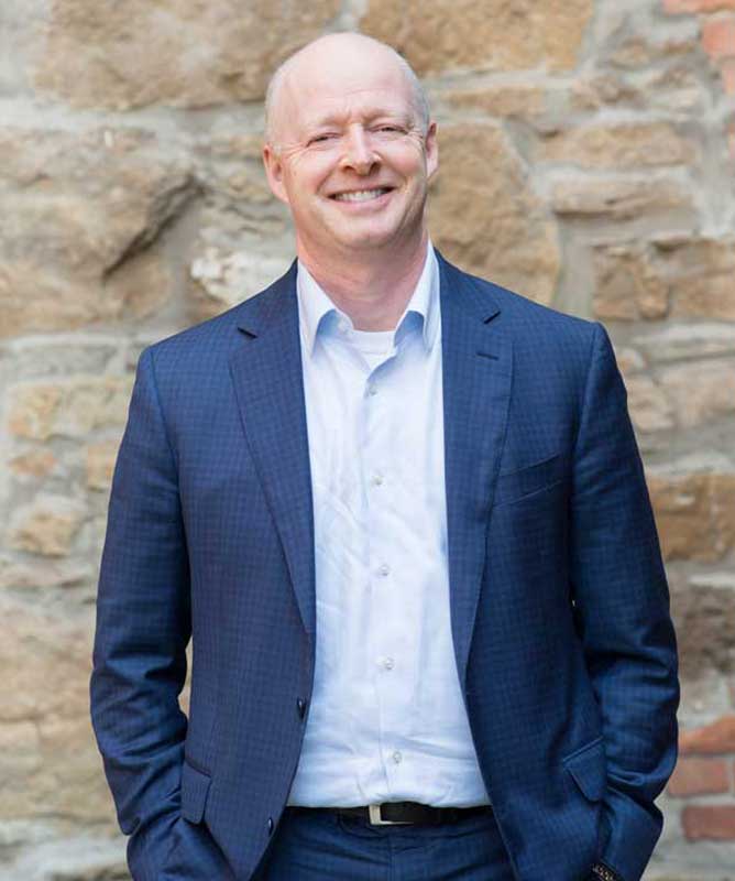 smiling bald man wearing nay suit and light blue shirt in front of stone wall