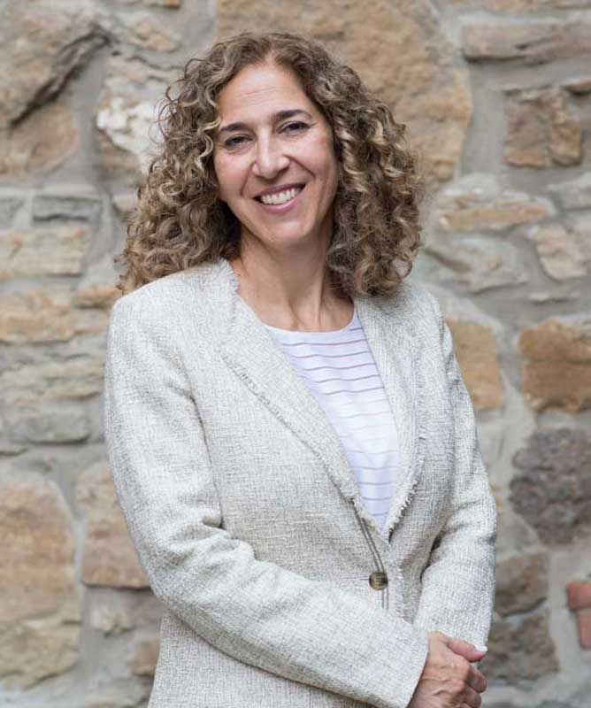 smiling woman with curly hair wearing light blazer and striped white shirt in front of stone wall