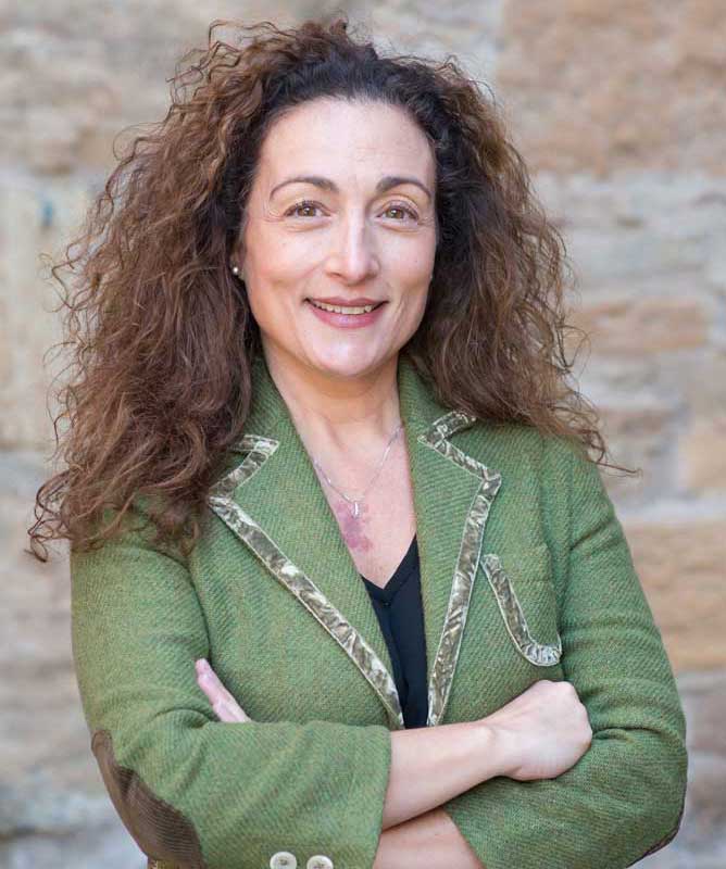 smiling woman with brown hair wearing green blazer with arms crossed in front of stone wall