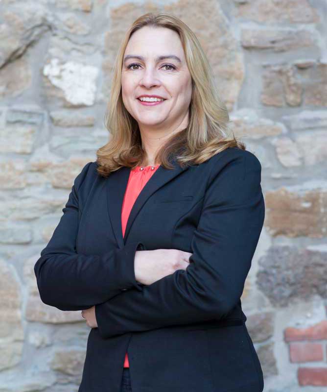 smiling woman with long blonde hair wearing navy blazer and red shirt with arms crossed in front of stone wall